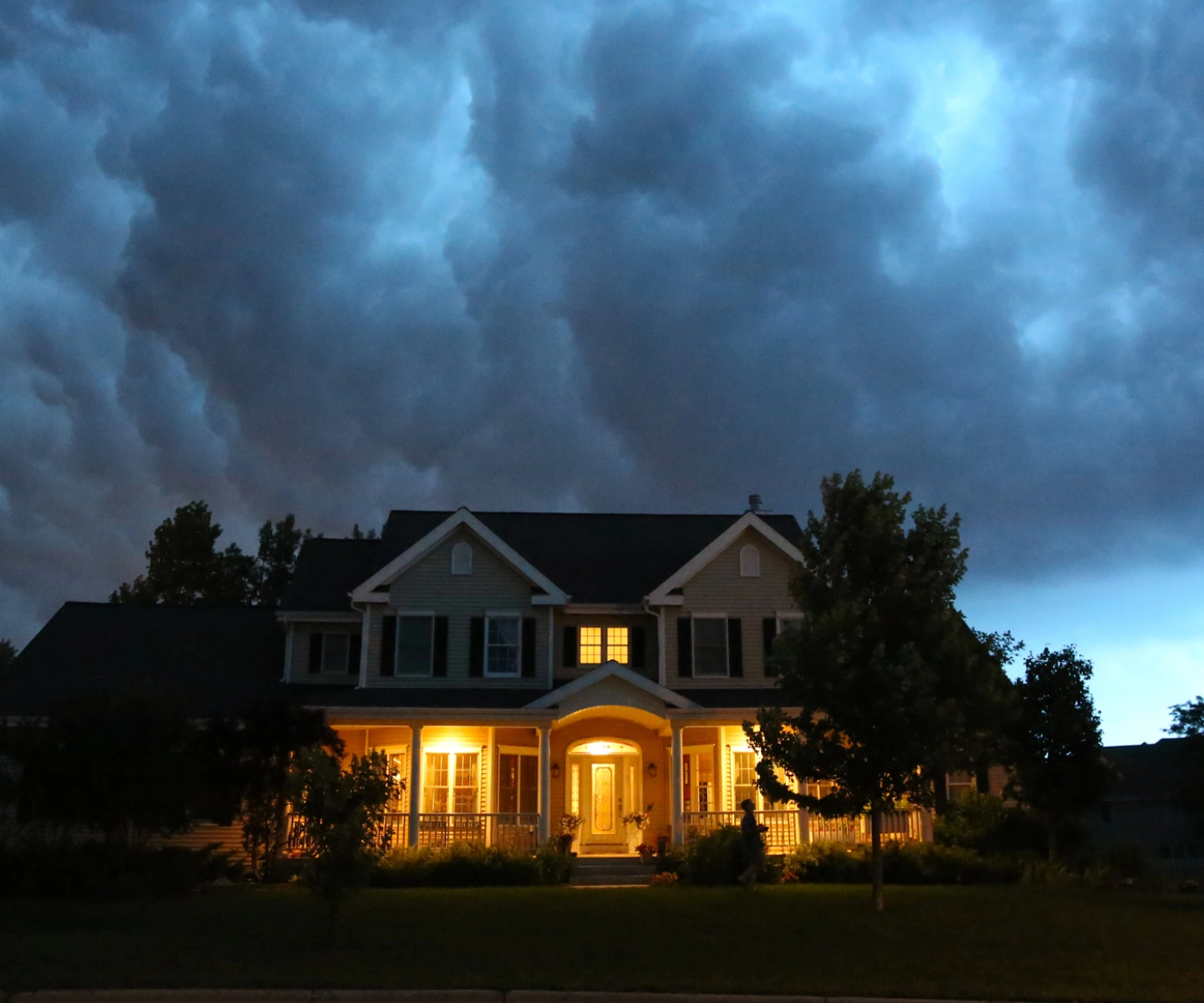 A two-story suburban home with its lights on as gray, stormy clouds gather behind it.