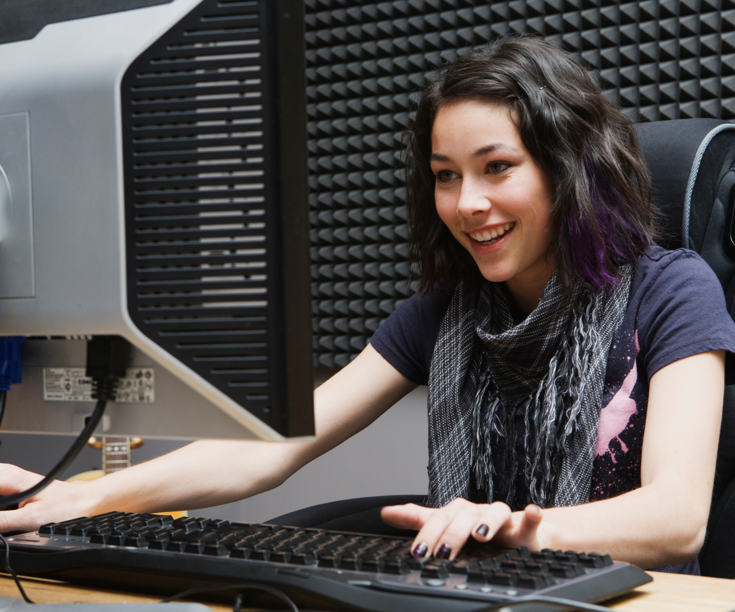 Woman in black t-shirt and gray scarf smiling while she plays on a desktop computer.