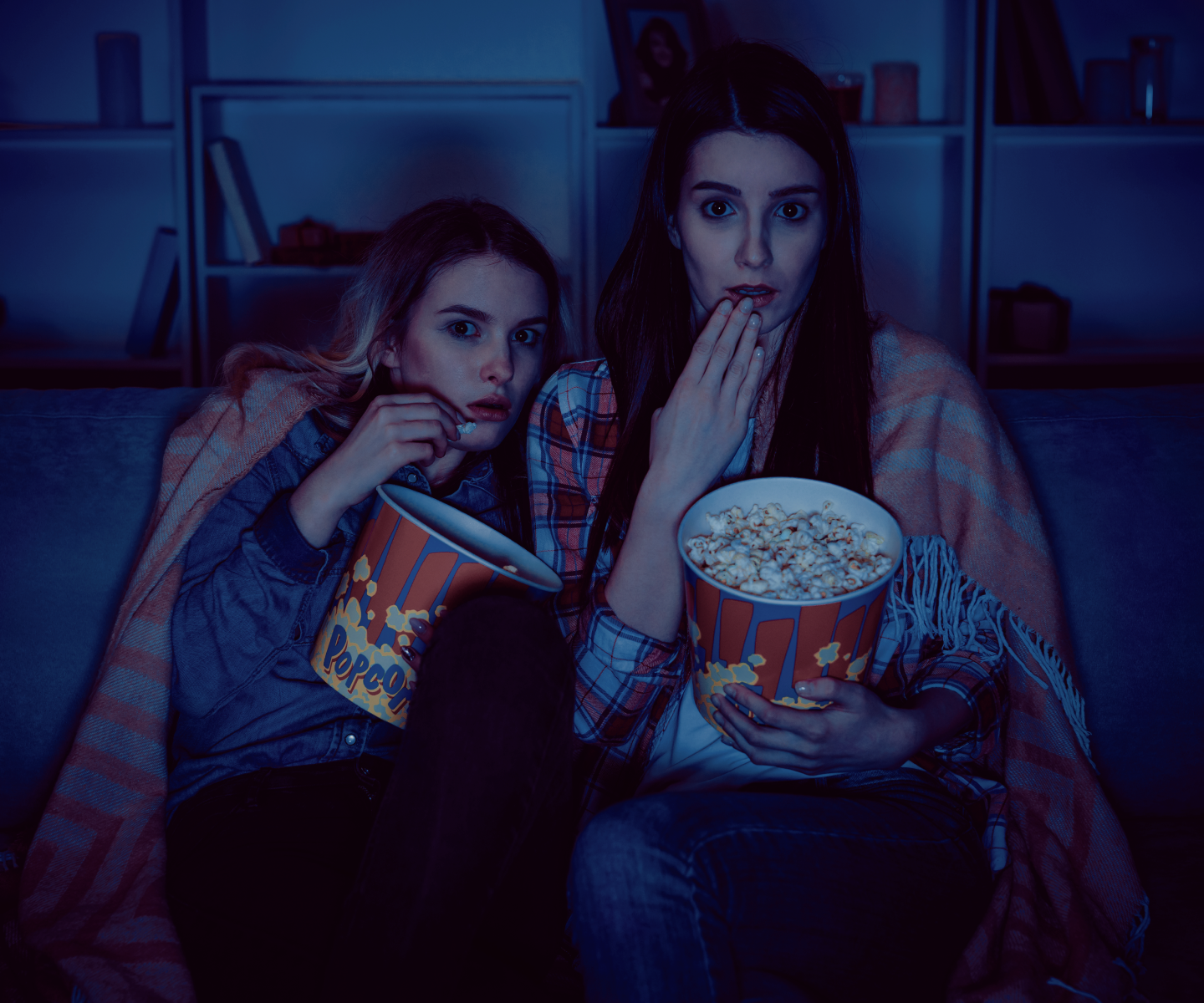 Two women are sitting with popcorn bowls in hand in the dark watching something on TV.