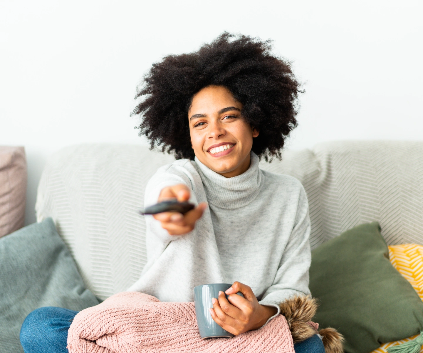 A woman sitting on a couch with a blanket and a coffee mug using the TV remote