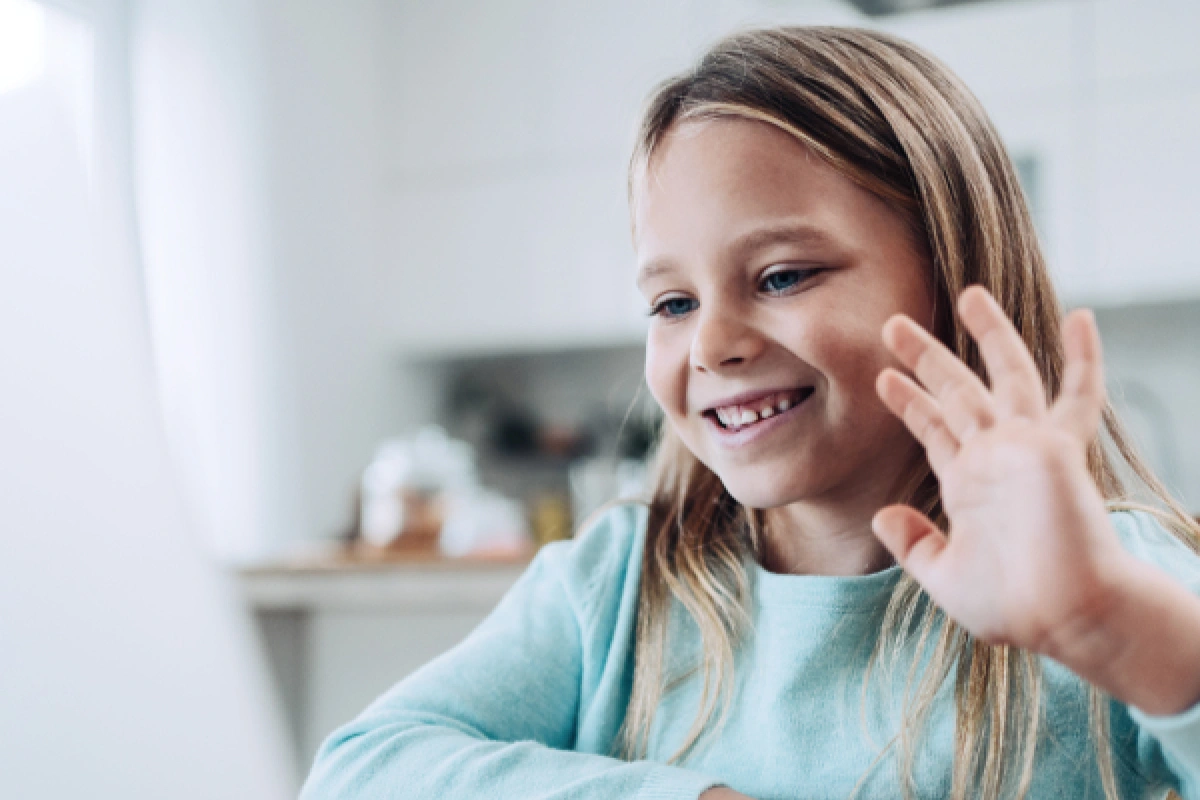 A smiling young girl sitting in front of a computer raising her hand as she participates in online school at home thanks to the Affordable Connectivity Program