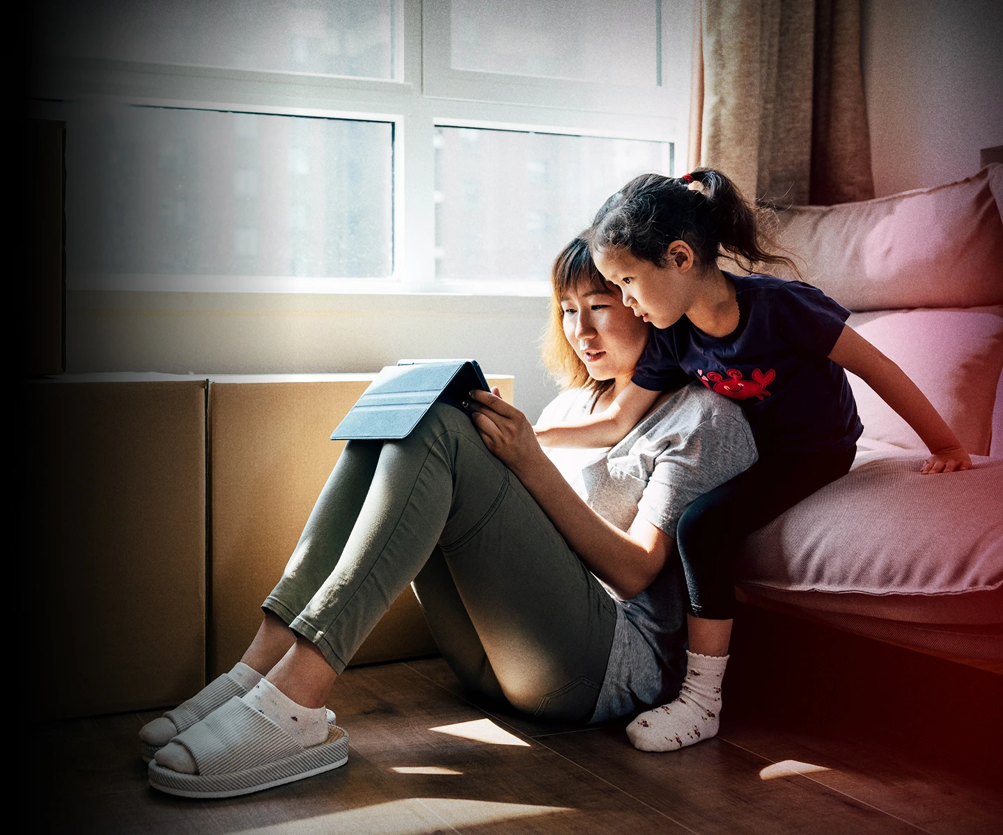 A mother and child sit on the floor among moving boxes, focused on a tablet together in their new home.
