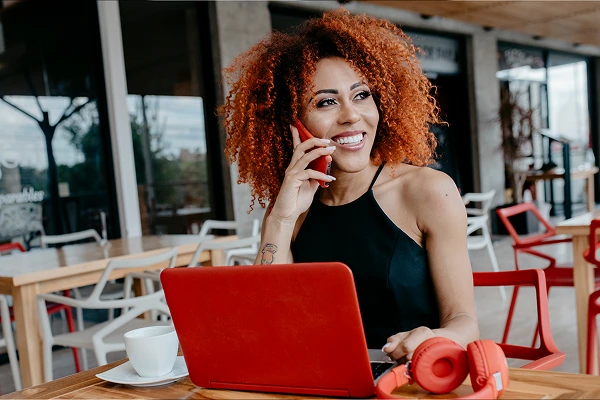 A smiling woman with curly red hair talking on a smartphone while working on a laptop at an outdoor cafe
