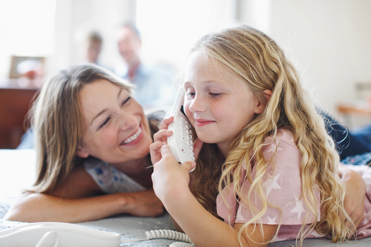 A young girl talking on a landline phone while her smiling mother watches.