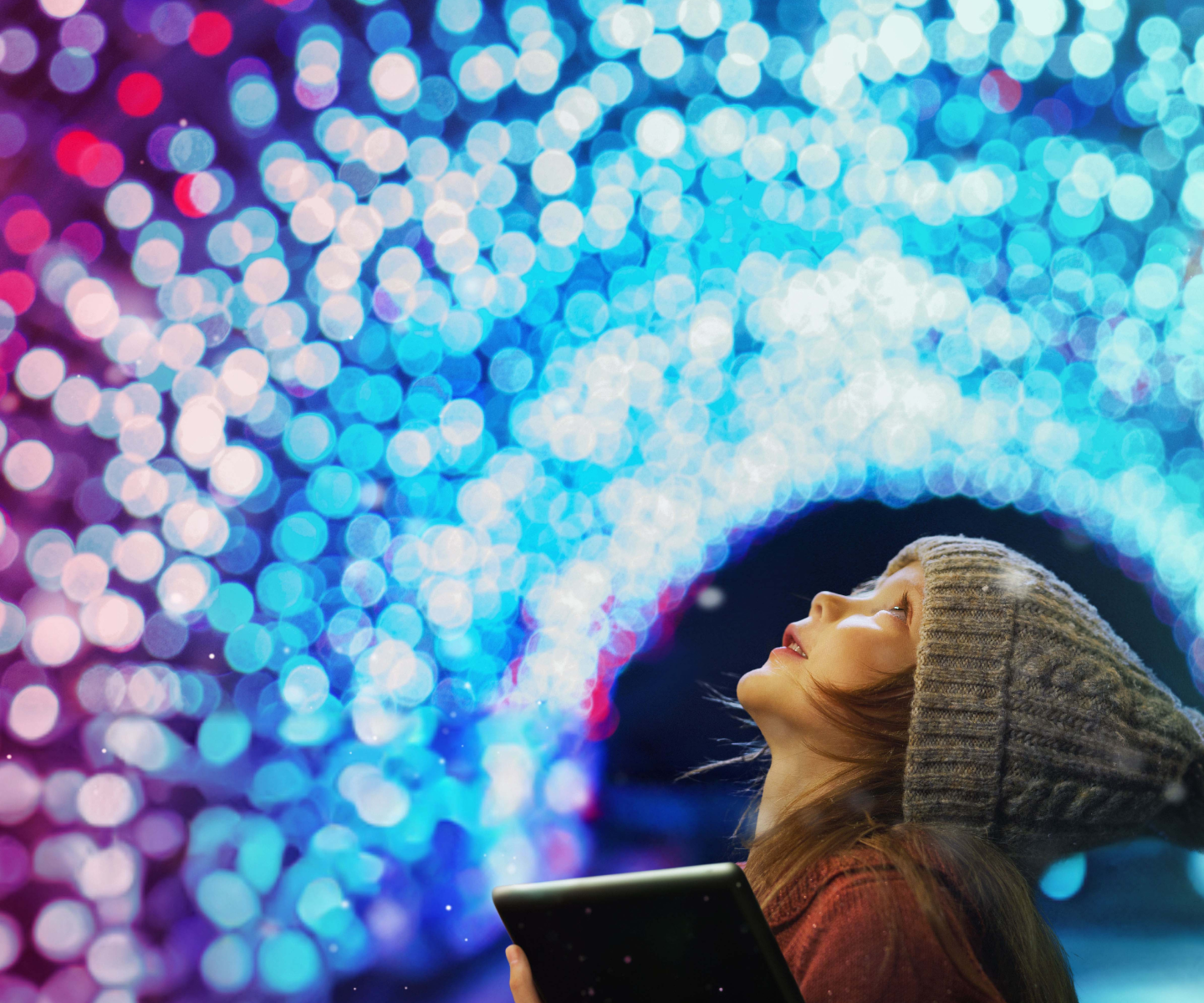 A child in a winter hat holding a tablet and staring at holiday lights.