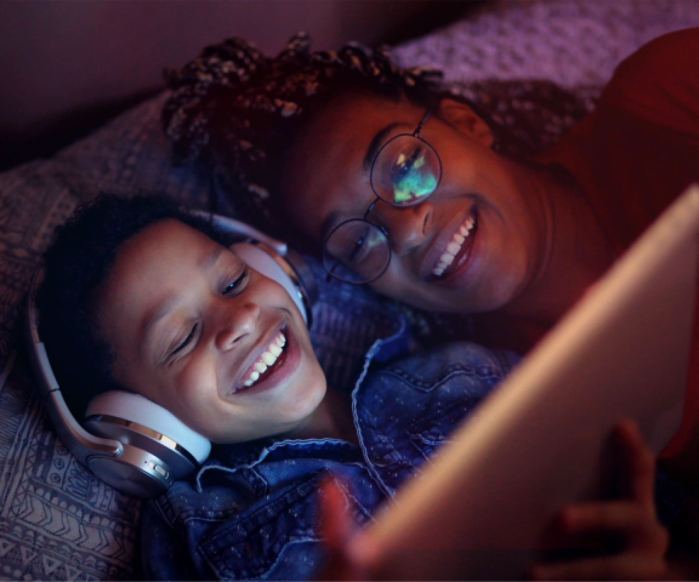 A child wearing headphones in bed and smiling alongside their mother while looking at a tablet