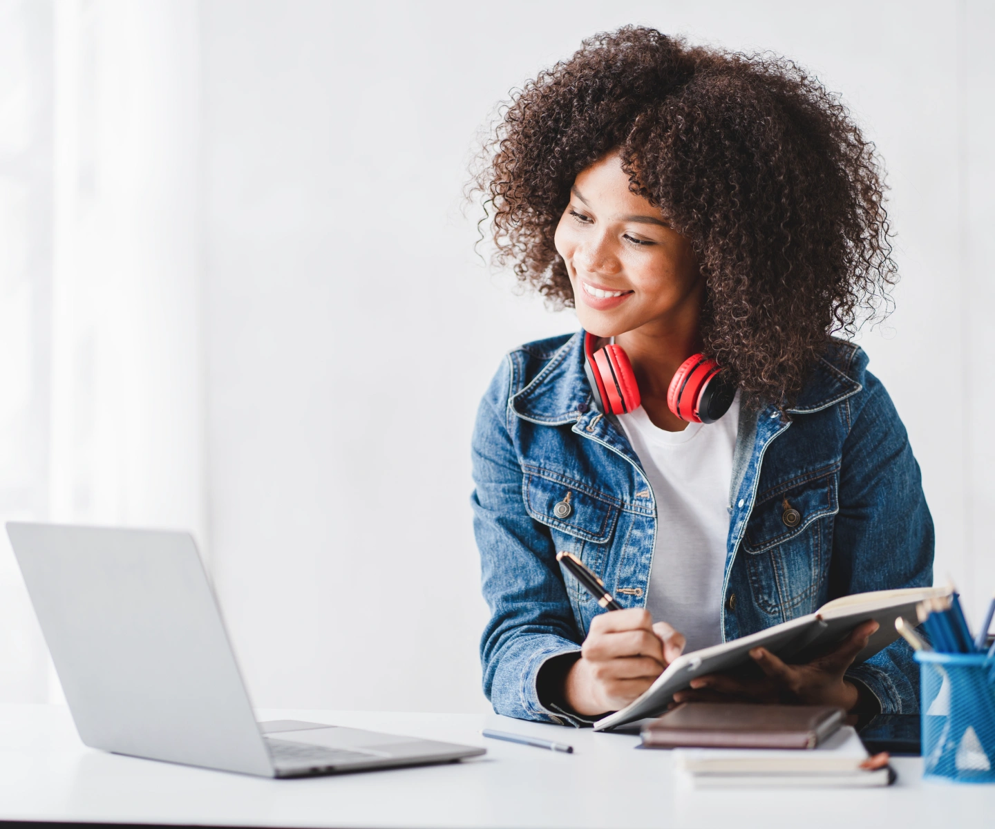 A woman with headphones around her neck writing into a notebook while looking at her laptop using Frontier Fiber