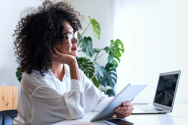 A woman sitting at a desk with multiple devices, including laptop, tablet and phone.