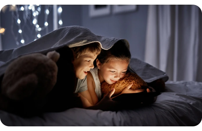 Two children look at a tablet under the covers in bed with their stuffed animals.
