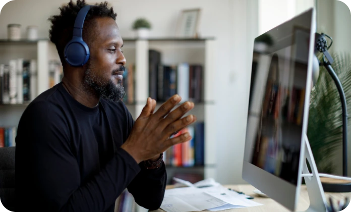 Man wears headphones and holds hands together while sitting at desk and looking at large computer screen