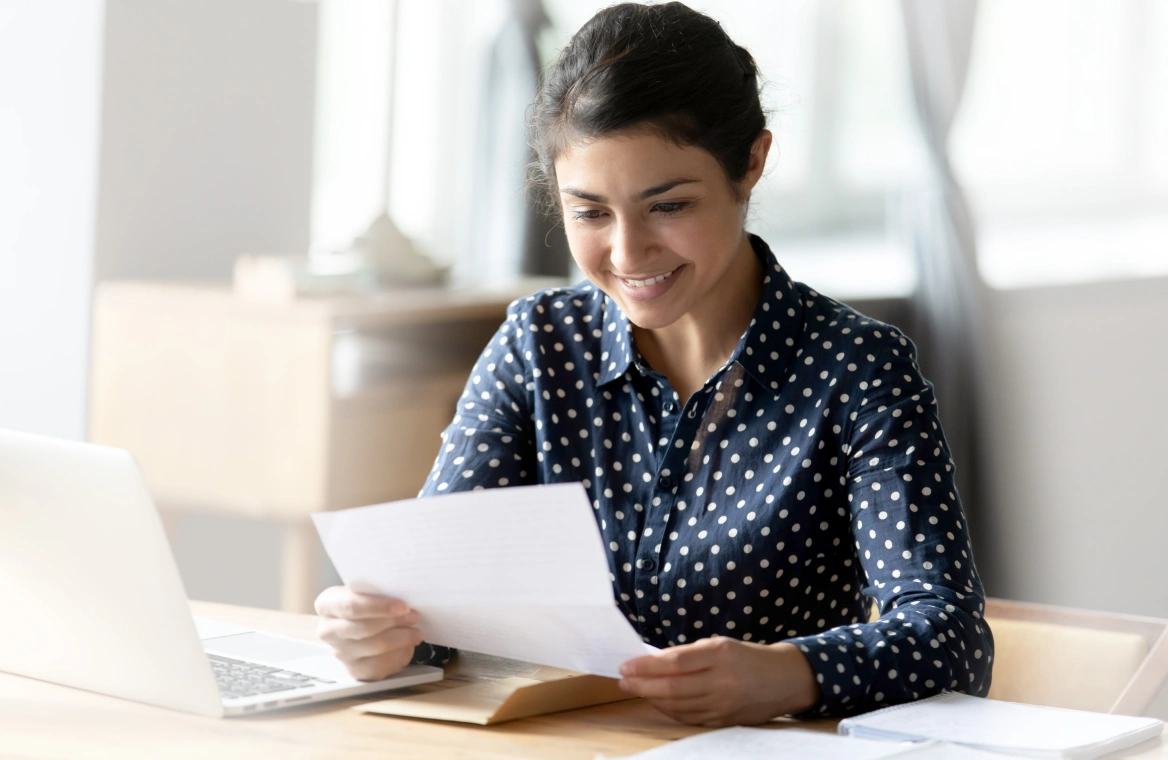 A woman sitting at her desk in front of a laptop and reviewing her paper bill