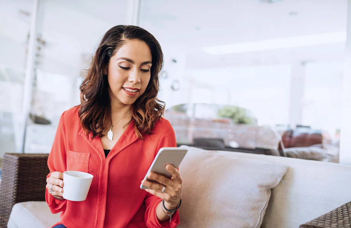 A woman sitting on the couch looking at her phone in one hand and with coffee in the other hand