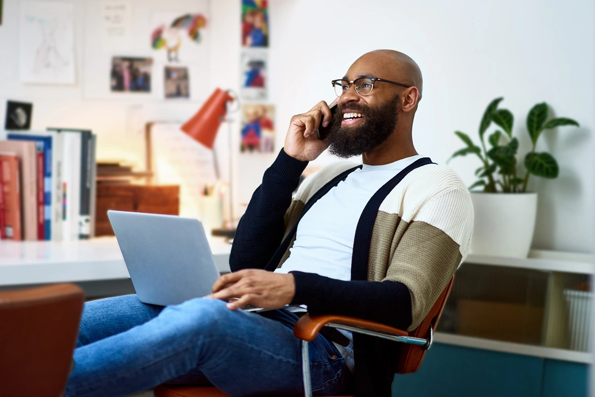 A smiling man talks on his phone while using a laptop at a home desk
