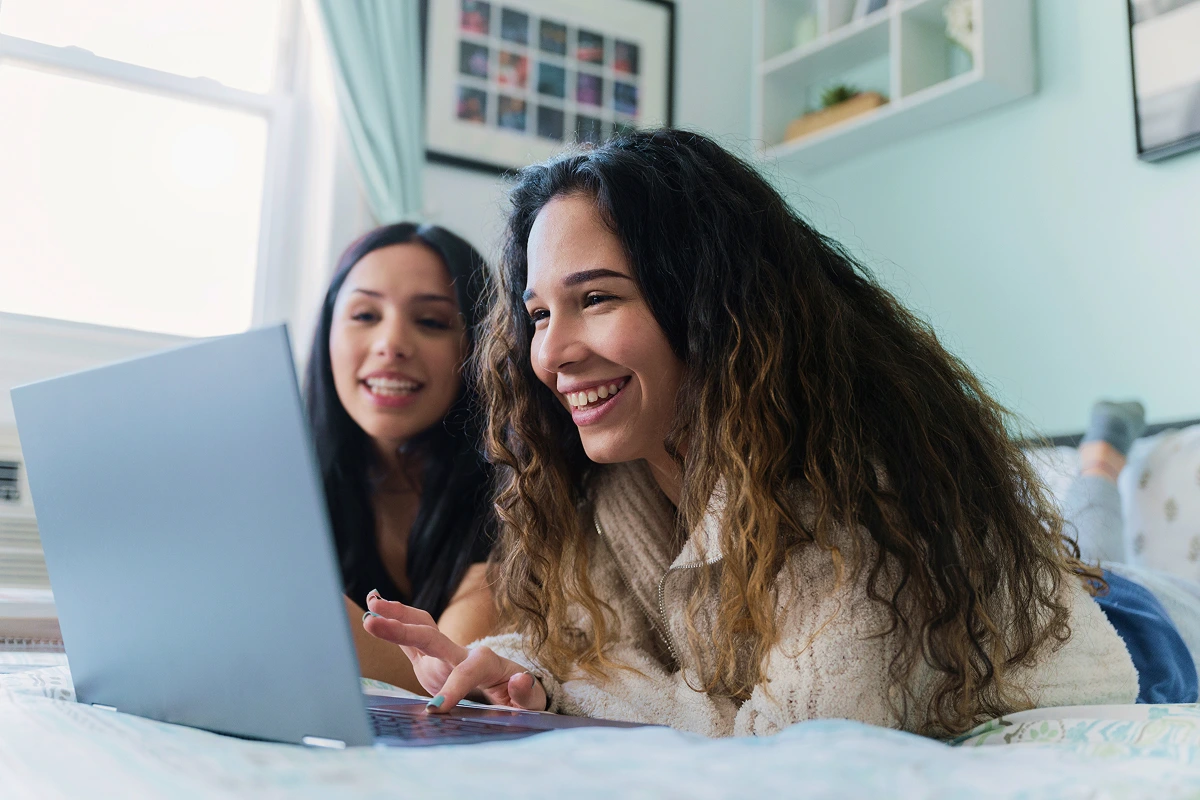 Two smiling young women laughing while browsing the internet together on a laptop in a bright bedroom