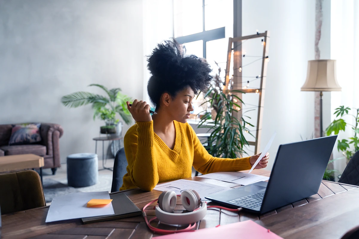A woman in a yellow sweater focuses on paperwork and a laptop while working from home in a modern living space